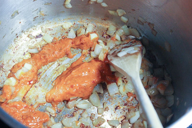Red curry paste and fried onions in metal pot, mixed by wooden spoon.