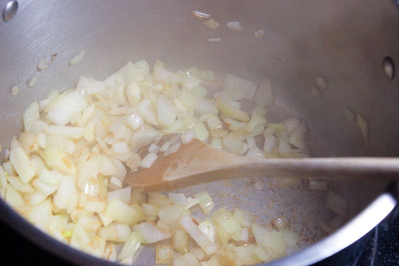 Onions frying in metal pot, mixed by wooden spoon.