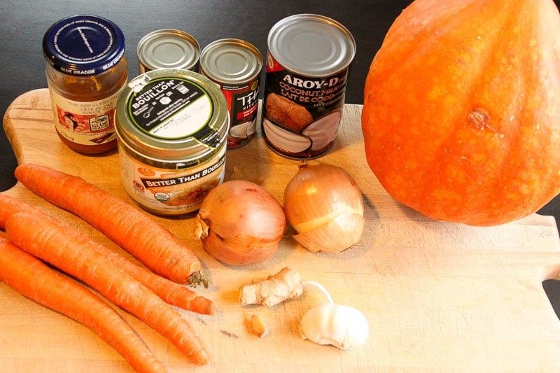 4 Carrots, jar of red curry paste, 2 small cans coconut milk, 1 large can of coconut milk, 1 squash, 1 jar of bouillon, 2 onions and a bulb of garlic on wood board.