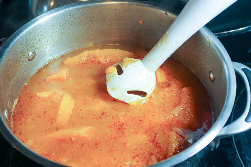 Immersion blender blending soup in metal pot on stove.