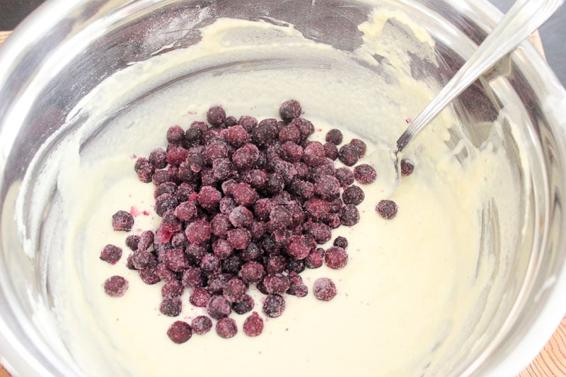 Frozen blueberries on top of pancake batter in metal mixing bowl.