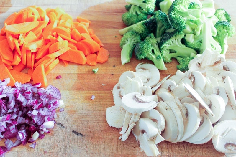 Piles of chopped red onion, carrot, broccoli and mushrooms on wood board.