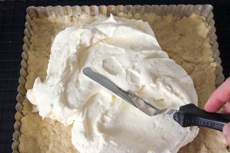 Spreading cream cheese on top of Shortbread crust in square tart pan.
