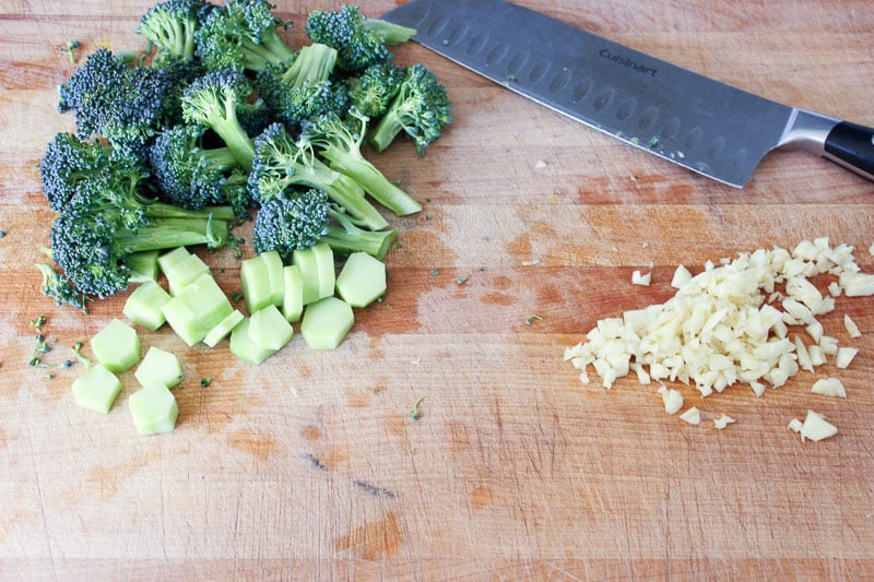 Chopped broccoli, minced garlic and large knife on wood board.
