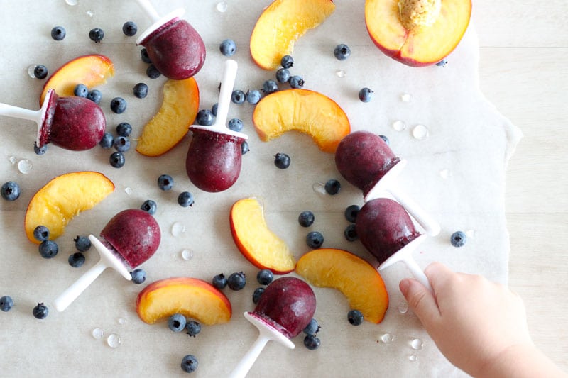 Child's hand picking up a Blueberry and Peach Popsicle with popsicles and fruit in background.