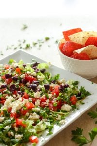 White square plate with hummus, tomatoes, parsley, feta and olives on it, with a white bowl of tortilla chips in the background.