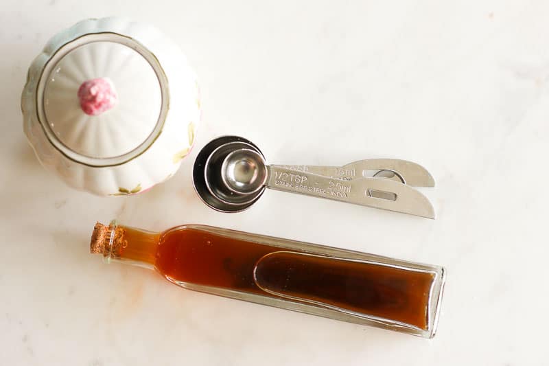 Narrow Glass Bottle of Vanilla Extract, White Sugar Dish and 3 Metal Measuring Spoons on White Board.