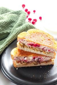 Two halves of a grilled sandwich with turkey, cream cheese, stuffing and cranberries, stacked one on top of the other, on a black plate. Green cloth and cranberries in the background.