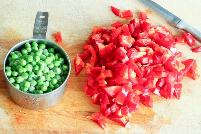 Metal Measuring Cup filled with Green Peas and Chopped Red Pepper on Wooden Board.
