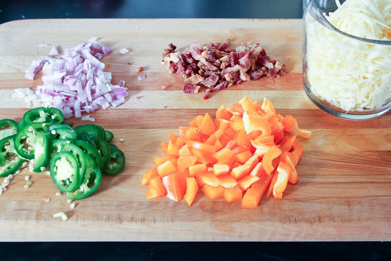 Shredded Mozzarella Cheese, Chopped Jalapeños, Bacon, Red Onion and Orange Peppers on Wooden Board.