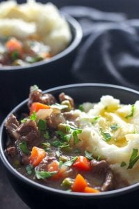 Steak and carrots in gravy with mashed potatoes in black bowl.