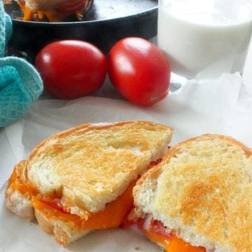 Grilled sandwich with cheese and tomato cut open to reveal melted cheese, with two tomatoes, a glass of milk and a cast iron frying pan holding another grilled cheese sandwich in the background.