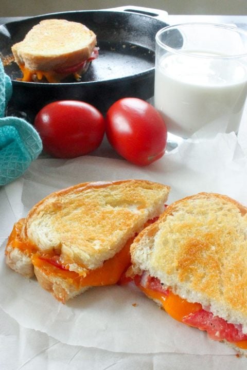 Grilled sandwich with cheese and tomato cut open to reveal melted cheese, with two tomatoes, a glass of milk and a cast iron frying pan holding another grilled cheese sandwich in the background.