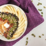 White bowl with yogurt, peanut butter, sliced banana, raisins, sunflower seeds and pumpkin seeds, with a purple cloth, with pumpkin seeds.