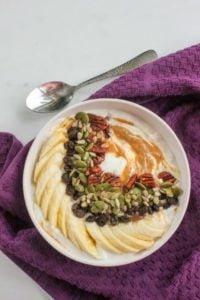 White bowl with yogurt, peanut butter, sliced banana, raisins, sunflower seeds and pumpkin seeds, with a purple cloth and a spoon.