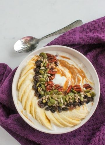 White bowl with yogurt, peanut butter, sliced banana, raisins, sunflower seeds and pumpkin seeds, with a purple cloth and a spoon.