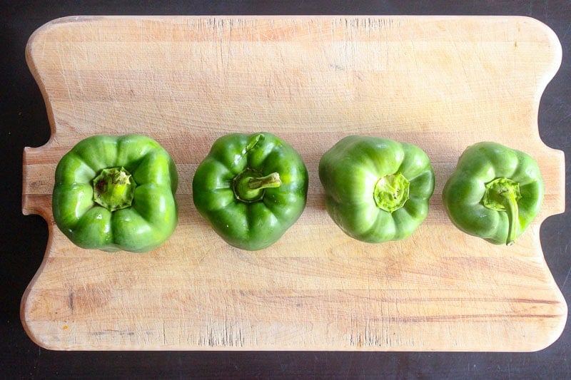 Four green bell peppers lined up on a Wooden cutting board.