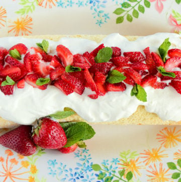 Cake roll covered with whipped cream and sliced strawberries, garnished with mint leaves, on a colourful platter decorated with flowers