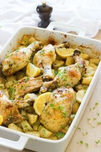 Pan of chicken and potatoes with lemon wedges and herbs in White Baking Dish.