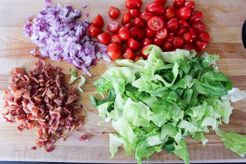 Diced red onion, halved cherry tomatoes, crumbled bacon and chopped romaine lettuce on wooden board.