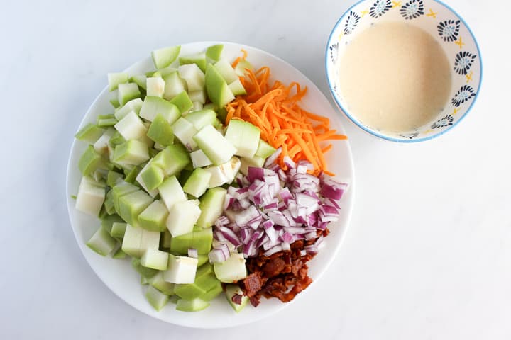 Plate of ingredients for chayote salad - cubed chaytote squash, minced red onion, crumbled bacon and shredded cheddar cheese. Dressing in bowl