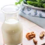 Carafe of creamy maple salad dressing on a cutting board with walnuts and a plate of mixed greens