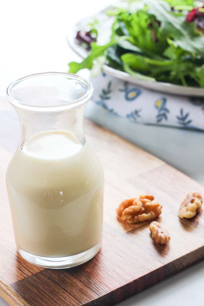 Carafe of creamy maple salad dressing on a cutting board with walnuts and a plate of mixed greens