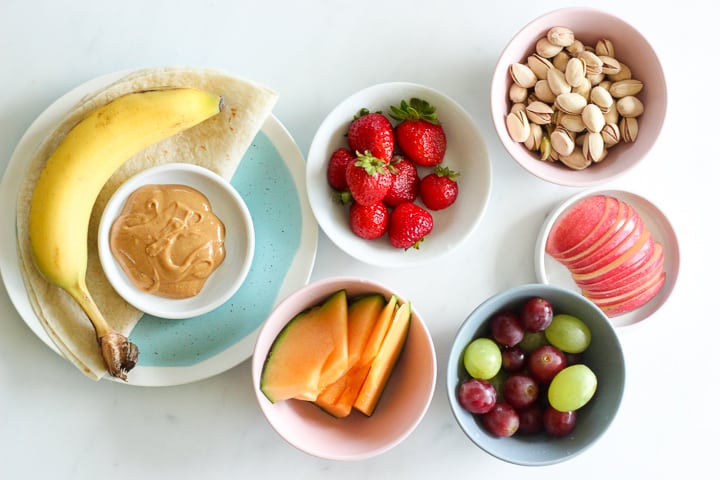 Plate with banana, tortilla and peanut butter, dishes holding strawberries, pistachios, cantaloupe, grapes and apple