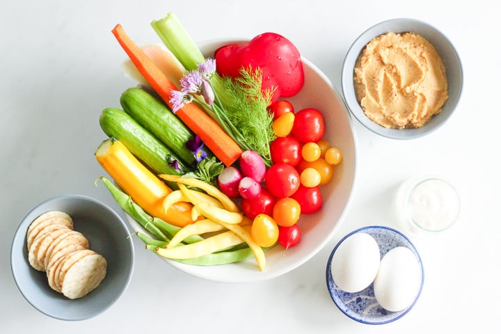 Bowl of fresh vegetables - cucumber, radishes, tomatoes, summer squash, green and yellow beans, dish of hummus, hard boiled eggs and rice crackers