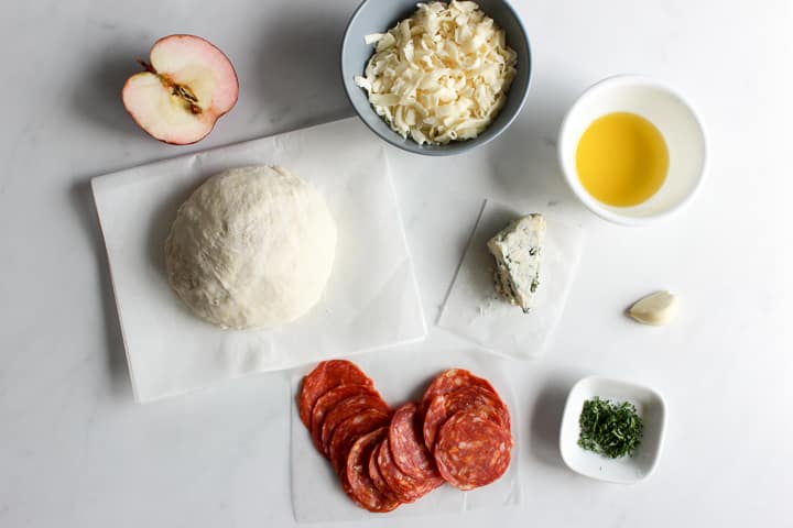 Pizza toppings including ball of dough, half an apple, grated mozzarella cheese, wedge of blue cheese, dish of olive oil, garlic clove, minced parsley and spicy salami