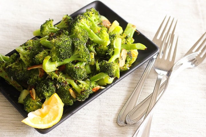 Black plate piled with roasted broccoli and lemon wedge