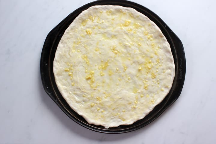 Thin crust pizza dough spread in round pizza pan with garlic oil on it