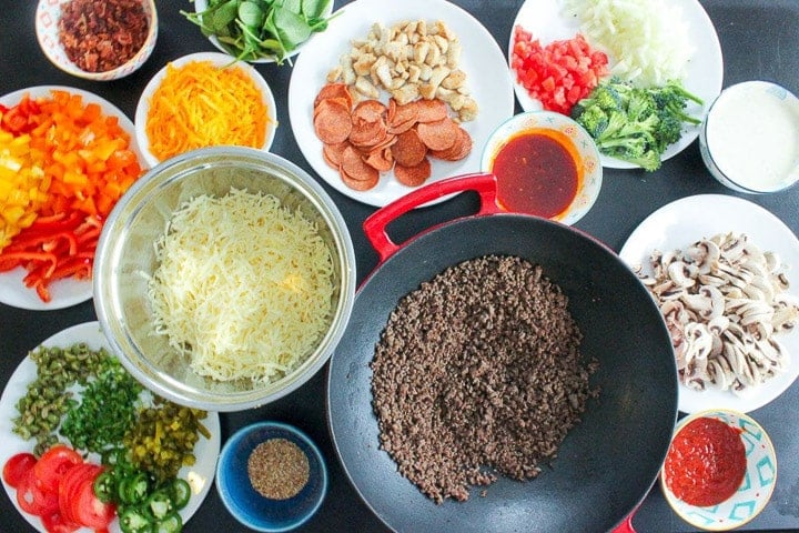 Overhead view of pizza toppings including ground beef, shredded cheese, mushrooms, peppers, vegetables and sauce