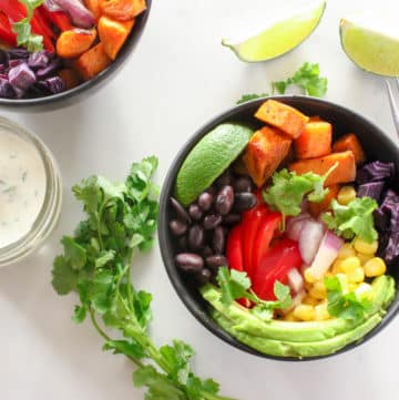 Two black bowls of roasted sweet potato cubes, sliced red peppers and avocados, black beans, corn kernels, cilantro leaves and a lime wedge