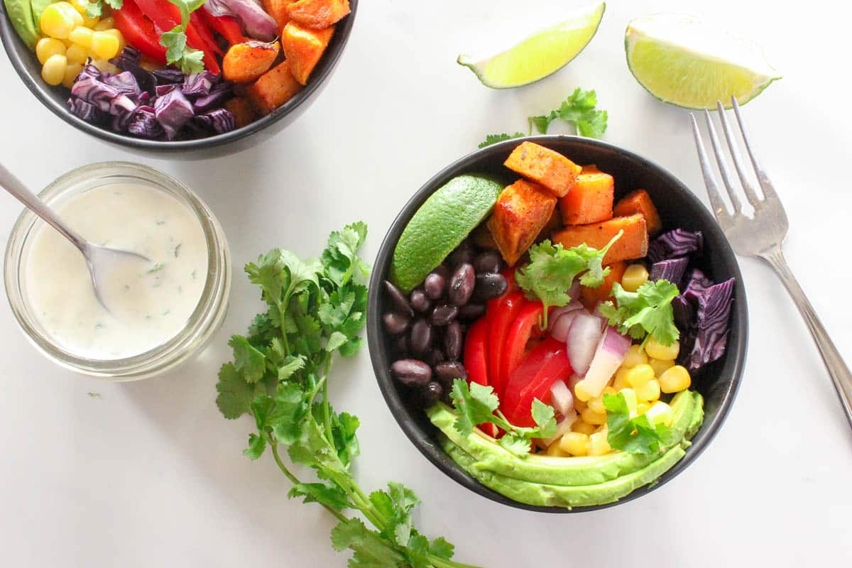 Two black bowls ofsweet potato, red peppers, avocado, black beans, corn, cilantro and lime 