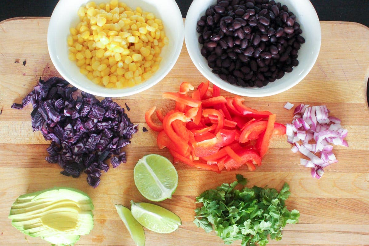 Cutting board with bowls of corn and black beans, red cabbage, red pepper, onion, avocado, lime and cilantro