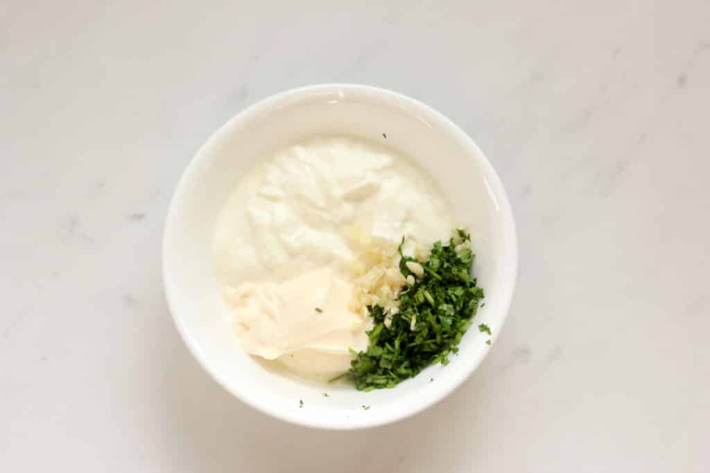 White bowl of yogurt, mayonnaise, chopped cilantro, minced garlic and lime juice on a white counter