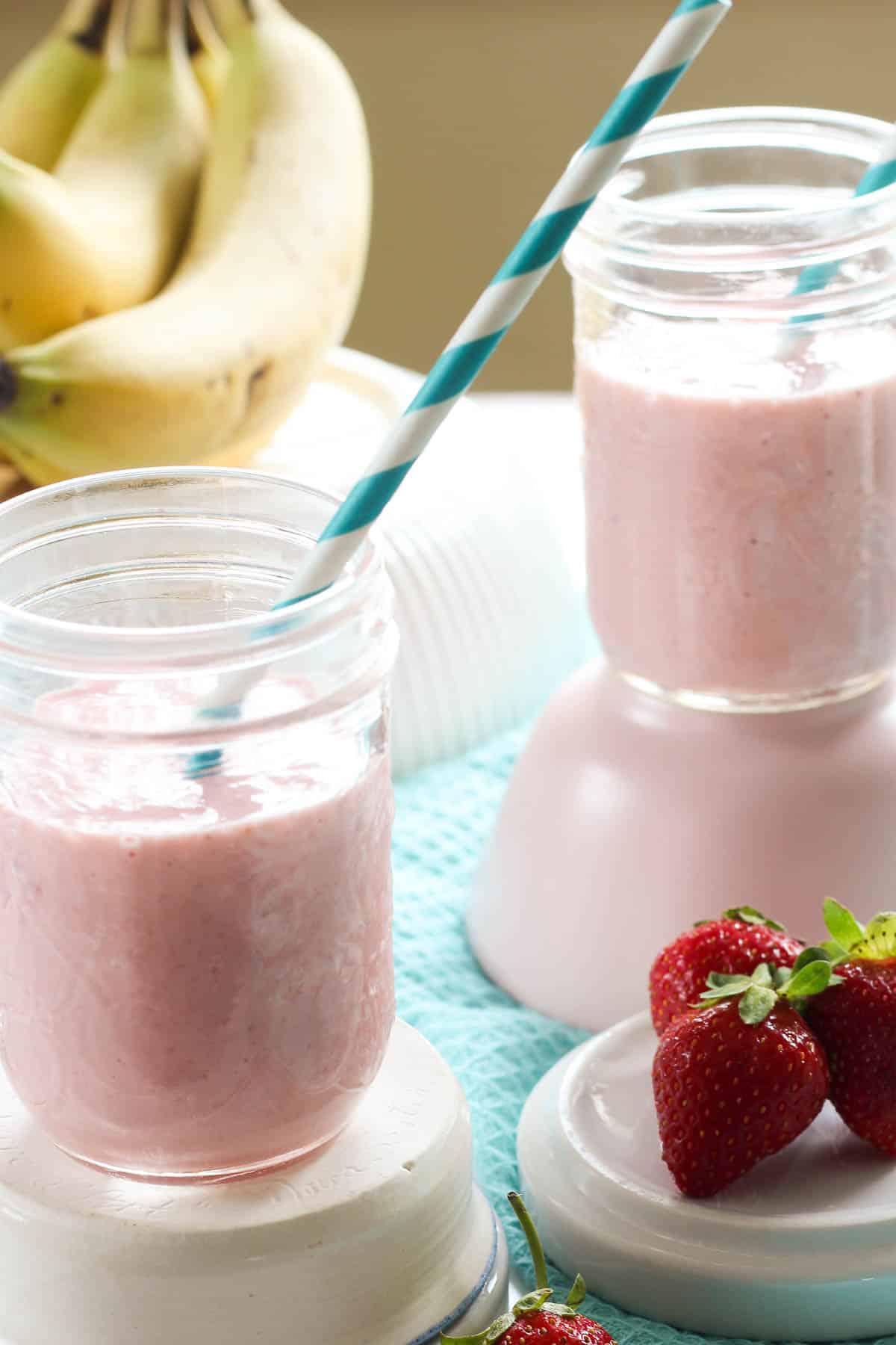 Two glasses of pink smoothie with blue and white strawbs, surrounded by strawberries and bananas and blue cloth
