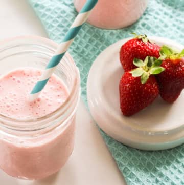 Glass of pink strawberry smoothie with blue and white straw and strawberries on a dish
