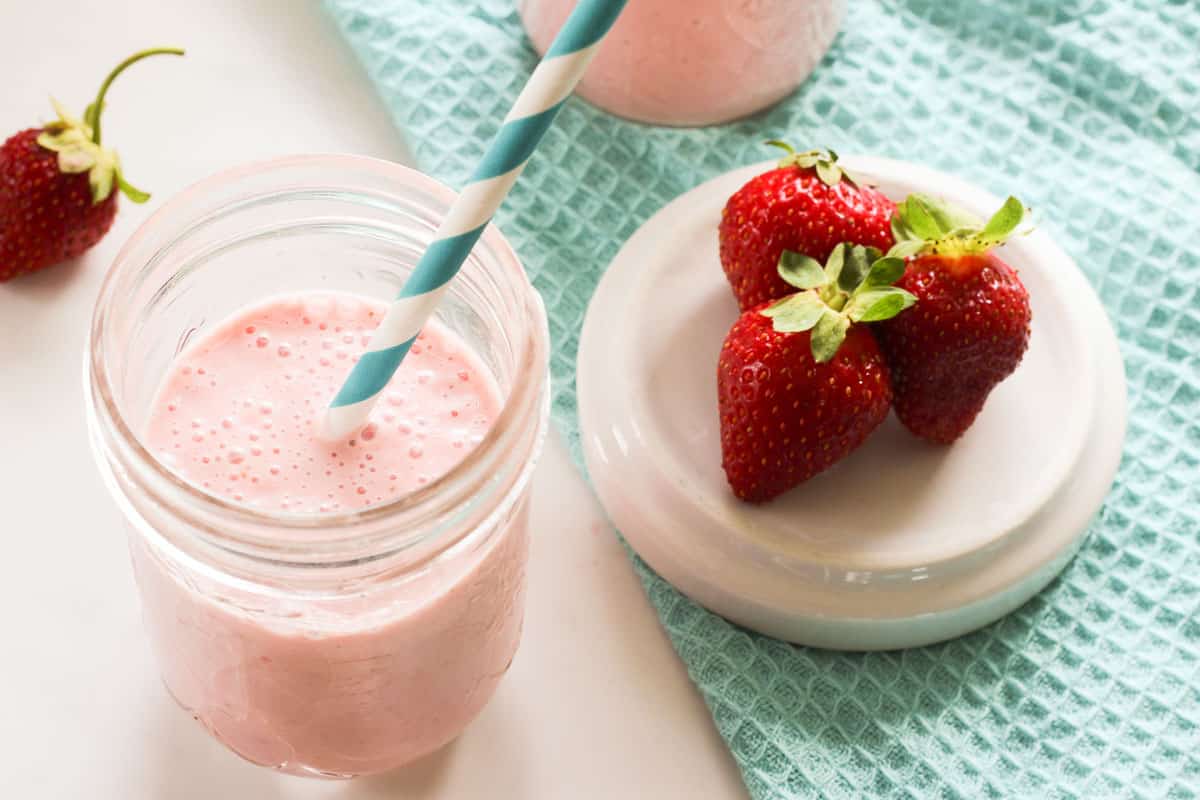 Glass of pink strawberry smoothie with blue and white straw and strawberries on a dish