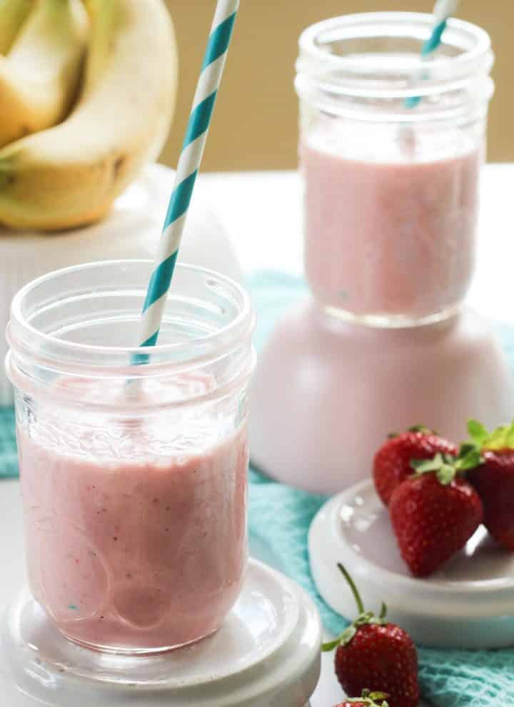 Two glasses of pink strawberry banana protein smoothie, with bananas and strawberries surrounding them and blue cloth.