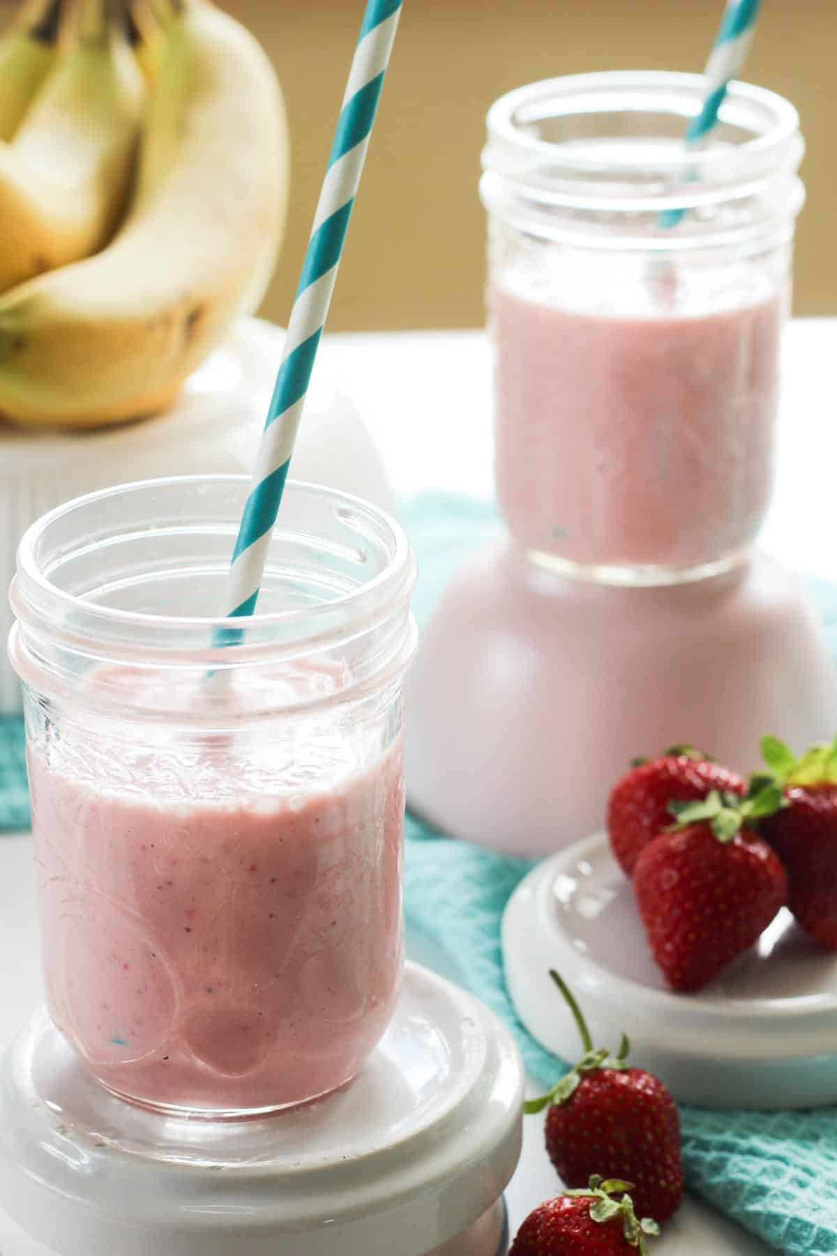 Two glasses of pink strawberry banana protein smoothie, with bananas and strawberries surrounding them and blue cloth.