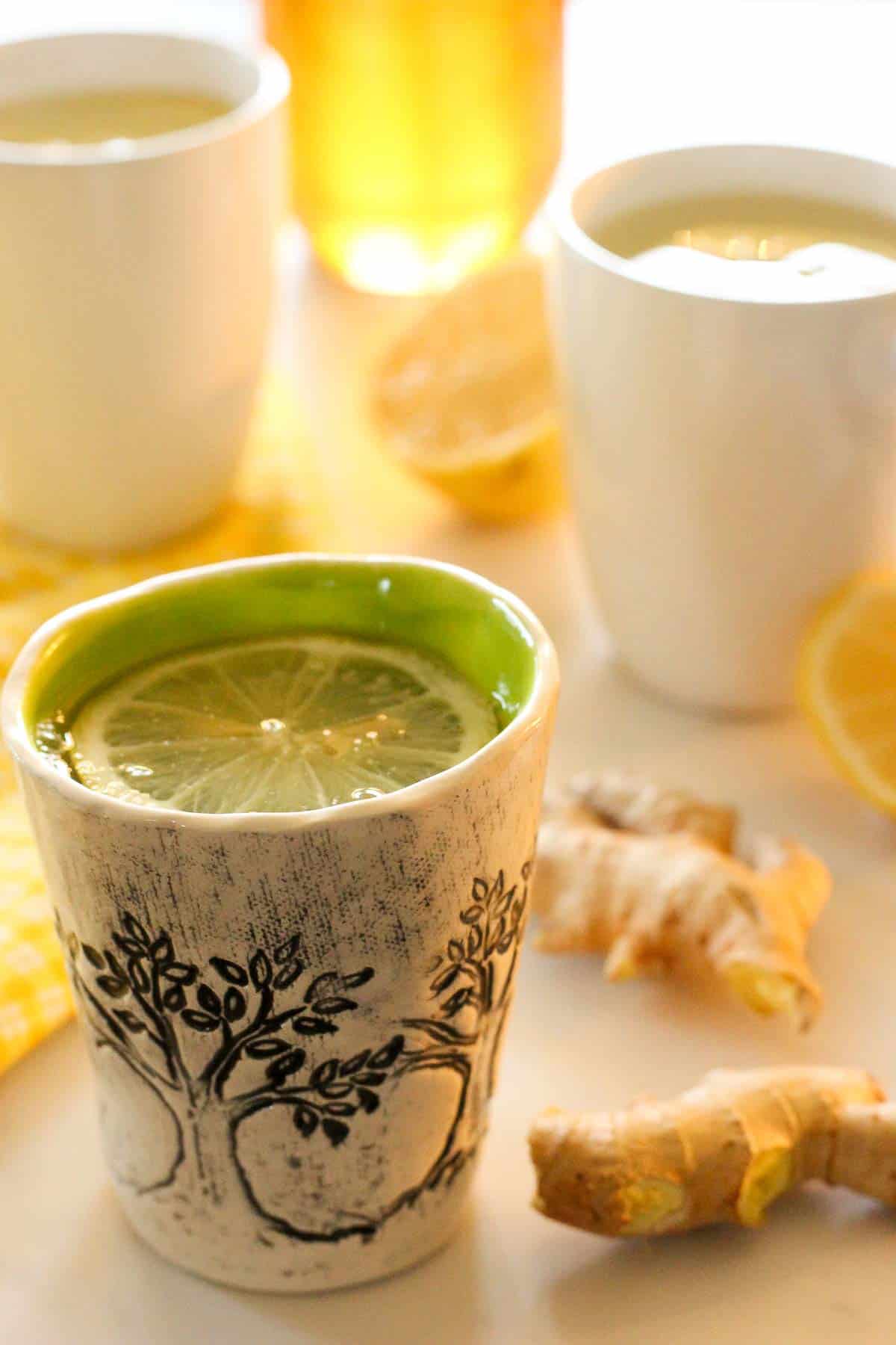 Pottery mugs of lemon ginger tea, with lemon slices, gingerroot and jar of honey in the background.