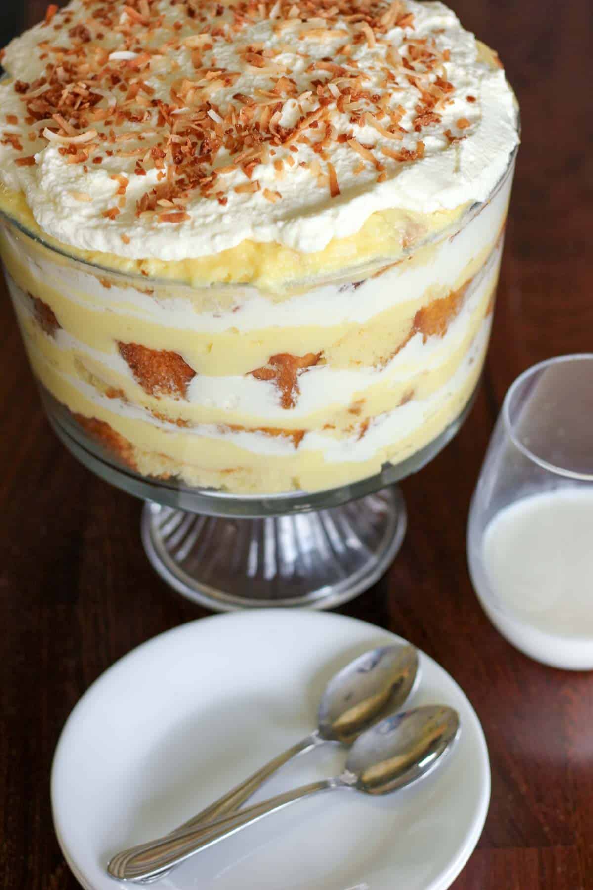 Dish of coconut cream trifle with plates, spoons and a glass of milk.
