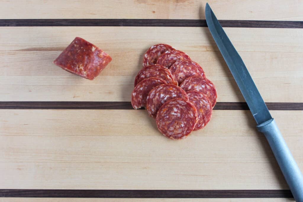 Sliced Genoa salami and knife on a wooden cutting board.