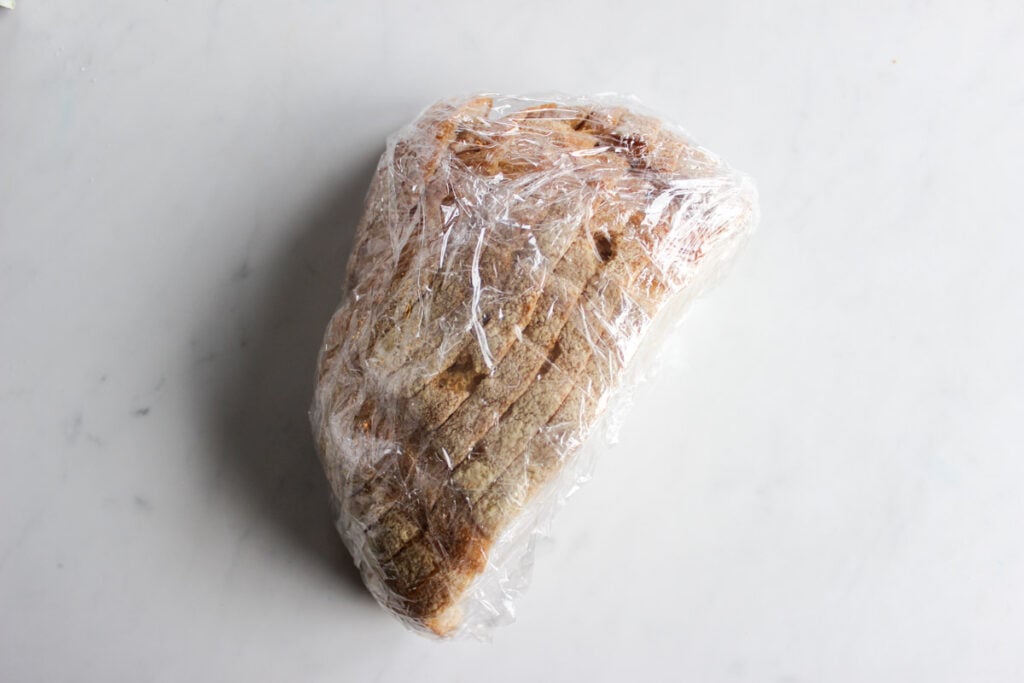 Half a loaf of sliced sourdough bread wrapped in plastic wrap.